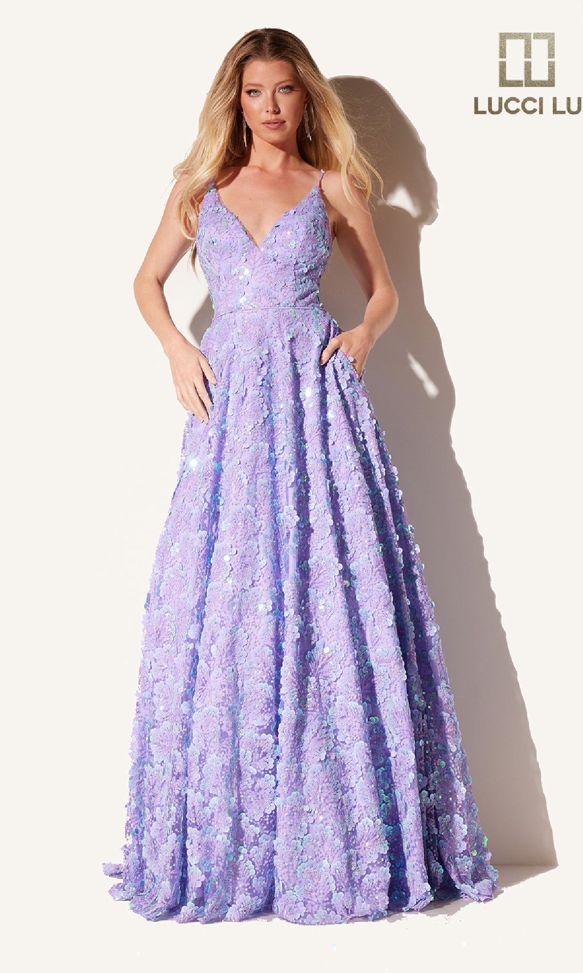 A woman wears the Long Prom Dress with Pockets: Lucci Lu 1325, a light purple, floor-length gown featuring a V-neckline and floral textured embellishments. The background is plain with the Lucci Lu logo in the top right corner.